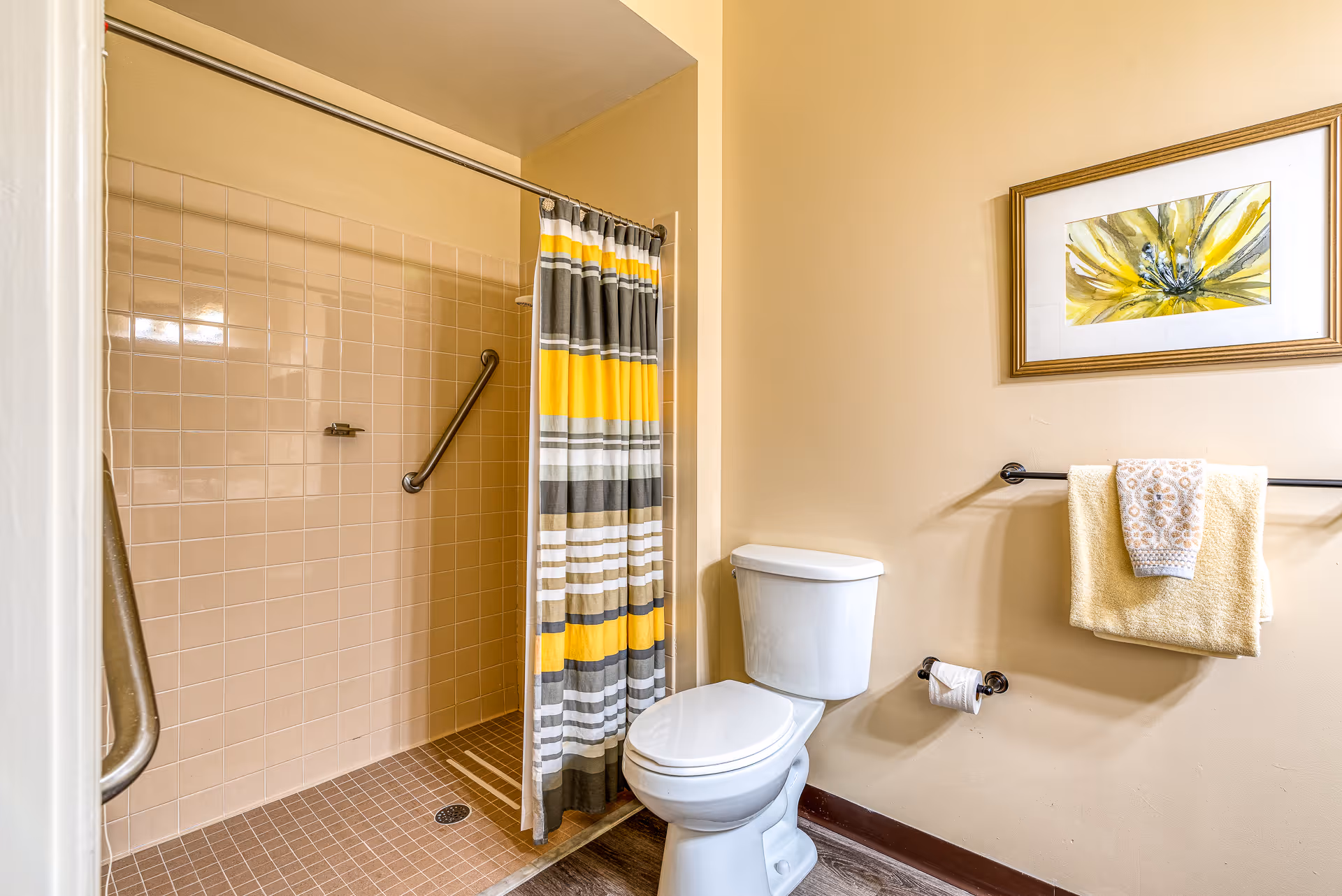 A bathroom with a white toilet next to a walk-in shower with beige tiles and a striped shower curtain in yellow, gray, and white. There is a towel rack on the wall holding a yellow towel and a smaller patterned towel, and a framed picture of a yellow flower hangs above the towel rack.