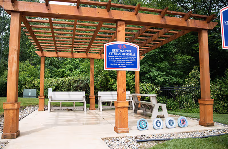 A wooden pergola structure with benches underneath, located outdoors in a green park area. A blue sign on the pergola reads 'Heritage Park Veteran Memorial.' Four circular military emblems are displayed on the ground in front of the pergola. Trees and bushes surround the area.