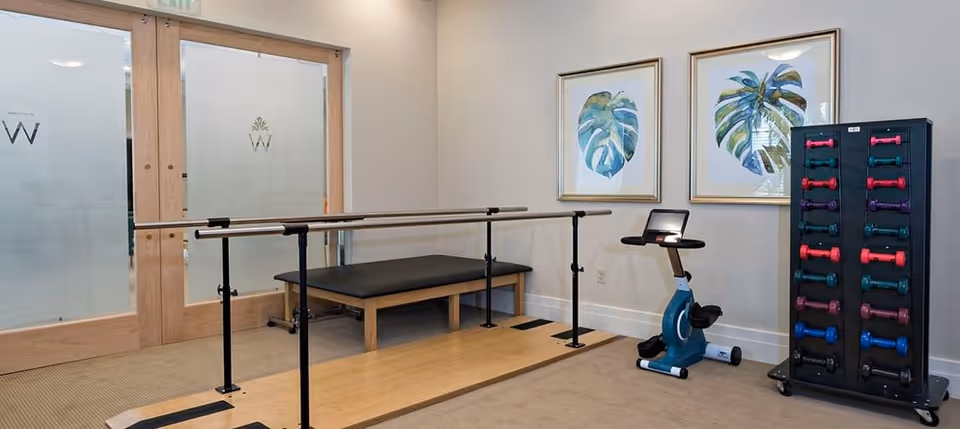 A small exercise room with parallel bars for walking support, a black padded bench, a stationary exercise bike, and a rack holding colorful dumbbells. The room has beige walls with two framed botanical prints and frosted glass double doors with wooden frames.