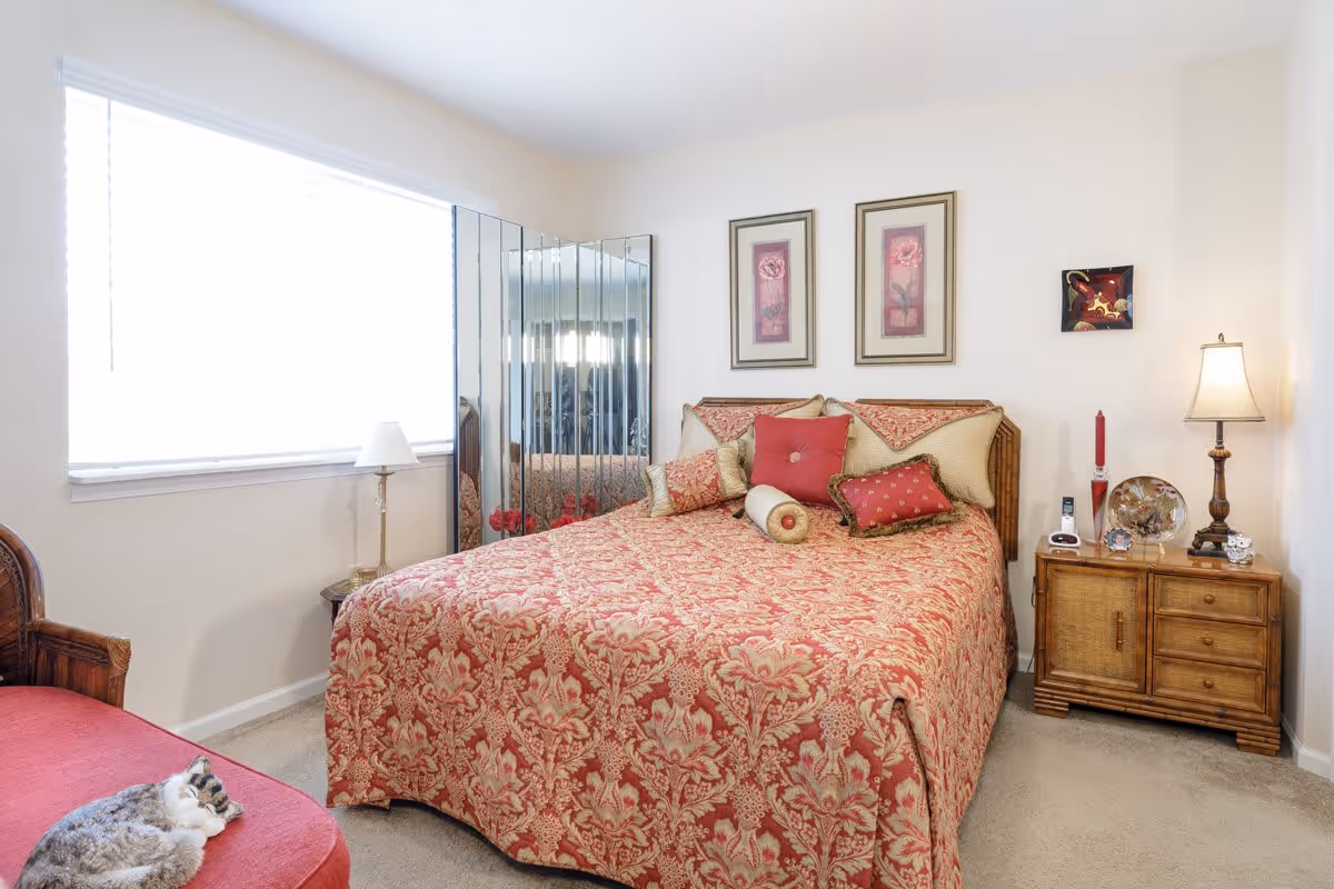 A cozy bedroom with a bed covered in a red and gold patterned bedspread and multiple decorative pillows. There is a wooden nightstand with a lamp, a red candle, and decorative items. A large window with blinds lets in natural light, and a cat is resting on a red cushioned chair in the corner. Two framed floral pictures hang above the bed, and a mirrored folding screen is positioned in the corner.