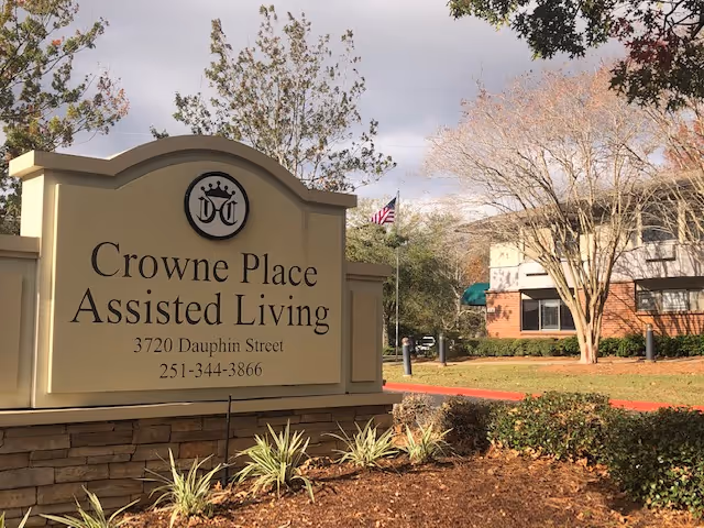 Outdoor view of the entrance sign for Crowne Place Assisted Living located at 3720 Dauphin Street with a building and trees in the background under a cloudy sky.