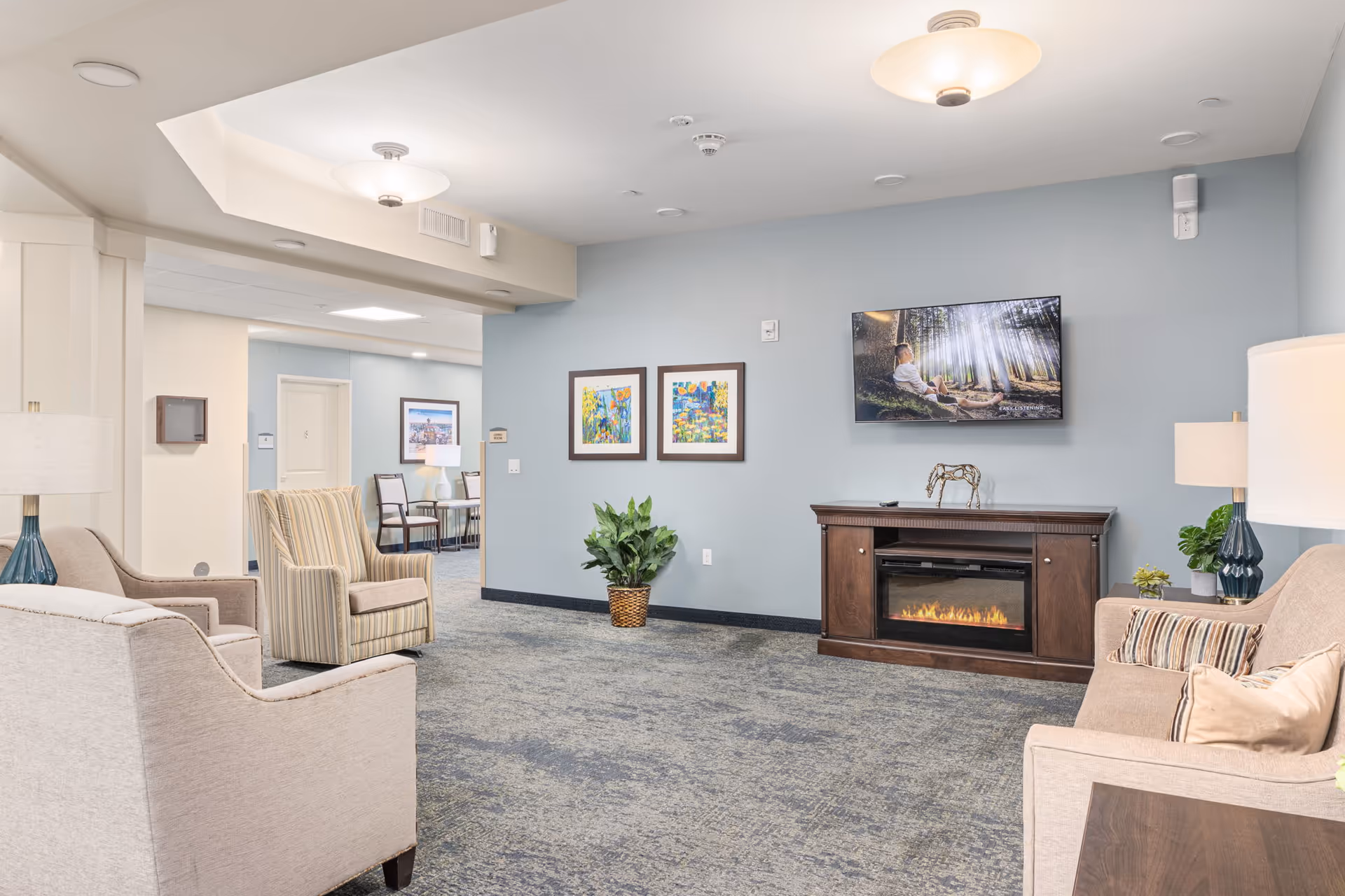 A cozy living room area in a senior living facility with beige armchairs and a sofa, a wooden electric fireplace with a TV mounted above it, two framed colorful paintings on a light blue wall, a potted plant, and two table lamps with blue bases.