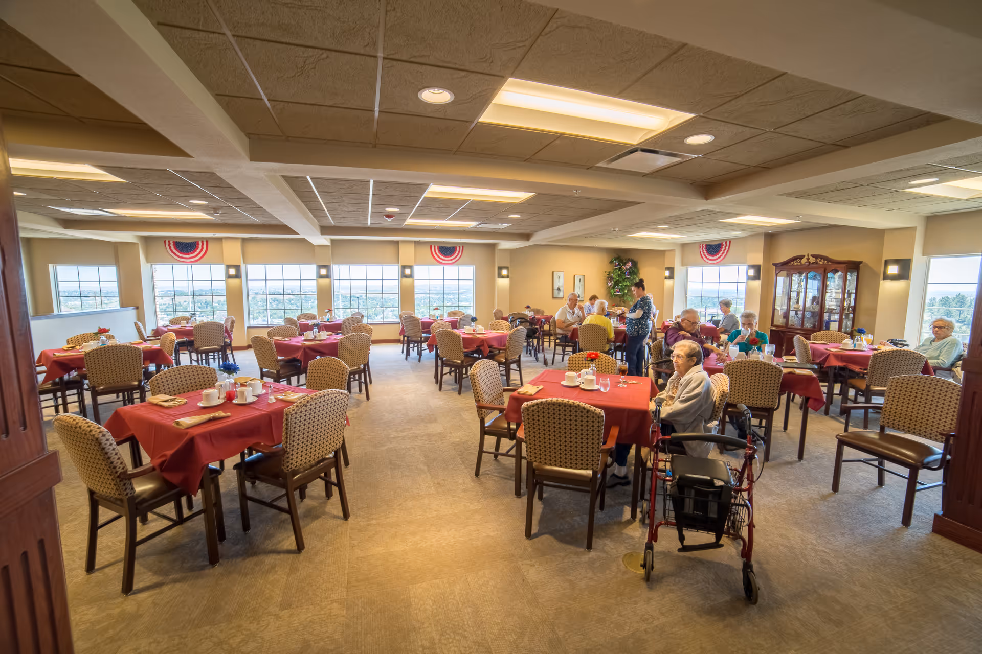 A spacious dining room in an assisted living facility with multiple tables covered in red tablecloths. Several elderly residents are seated at the tables, some using walkers or wheelchairs. Large windows provide a view of the outside, and the room is well-lit with ceiling lights. There is a wooden cabinet with glass doors on the right side and patriotic decorations hanging near the windows.