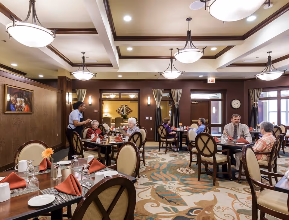 A dining room in a senior living facility with several elderly residents seated at tables, engaged in conversation. A staff member is attending to one of the tables. The room features elegant lighting fixtures, wooden paneling, and a patterned carpet. Tables are set with plates, glasses, and folded red napkins.