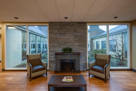 A cozy seating area with two brown armchairs facing each other on either side of a small wooden coffee table with magazines. Behind the chairs is a stone wall with an electric fireplace and a small plant on the mantel. Large windows on both sides of the fireplace provide a view of the building exterior and some greenery outside.