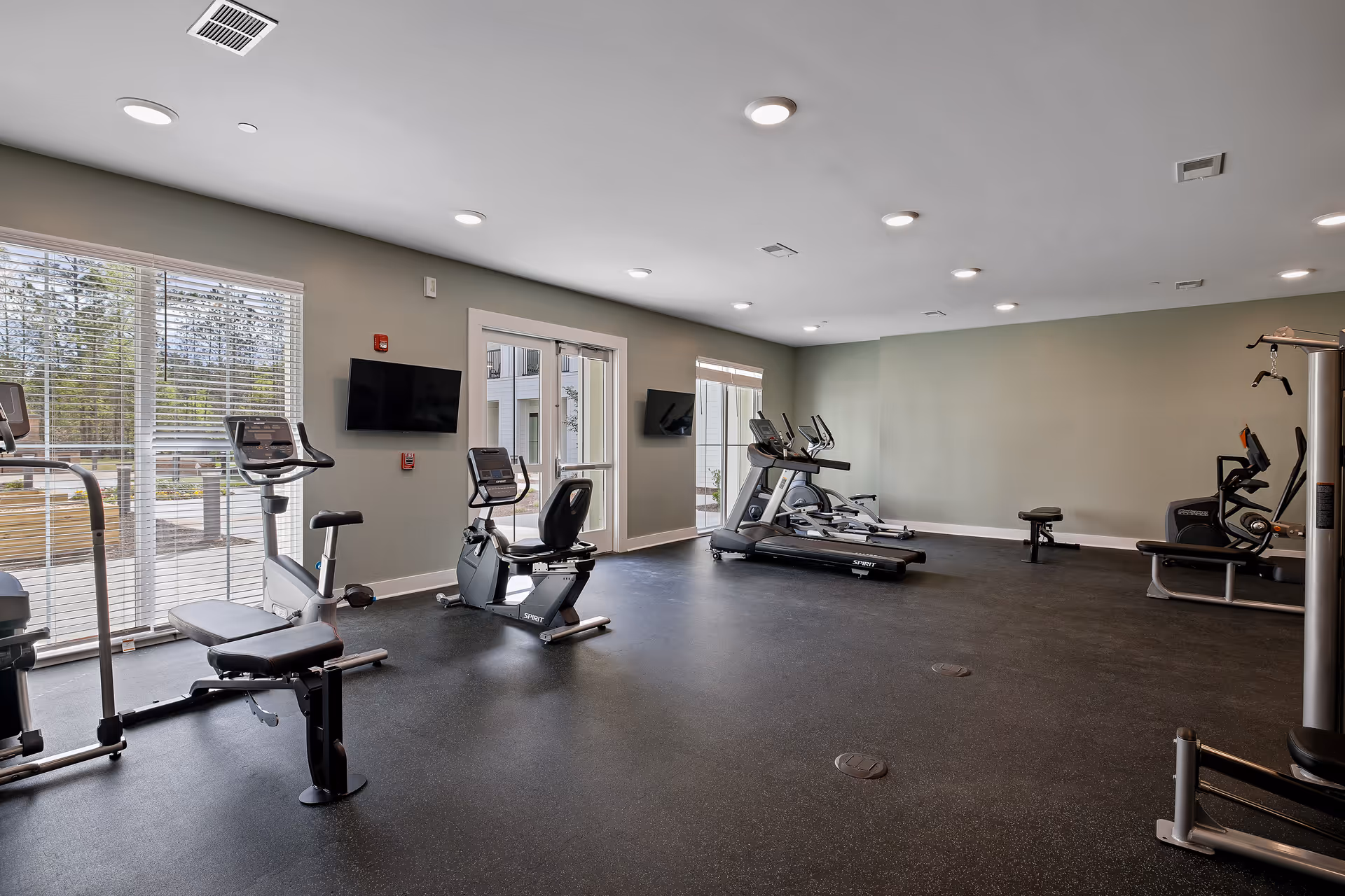 A spacious fitness room with exercise equipment including stationary bikes, treadmills, and weight machines. The room has large windows with blinds allowing natural light to enter, light gray walls, and recessed ceiling lights. Two wall-mounted TVs are visible, and there is a door leading outside.