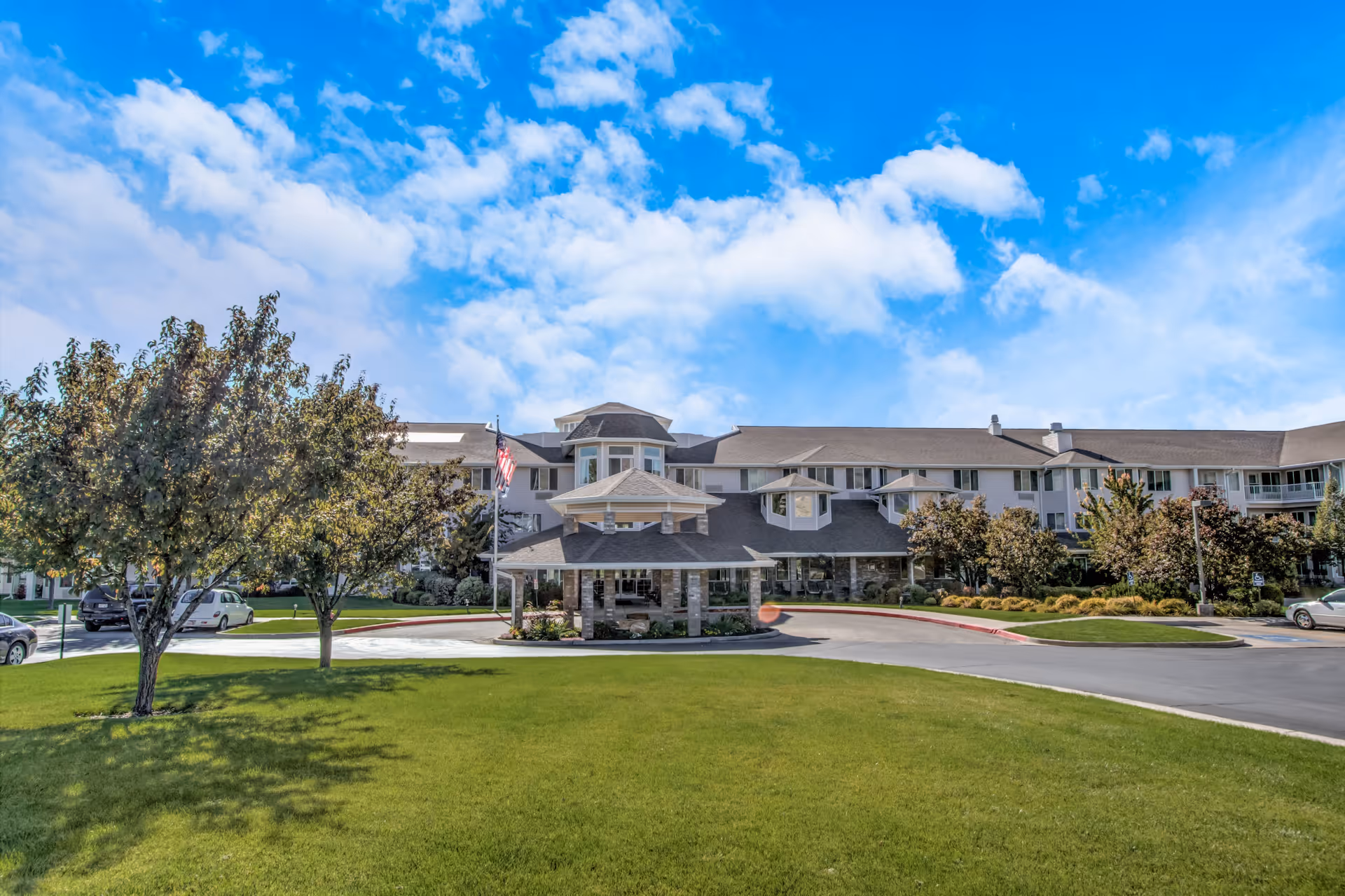 Front exterior view of Pioneer Valley Lodge Gracious Retirement Living building with a covered entrance, surrounded by green lawn, trees, and a clear blue sky with some clouds.
