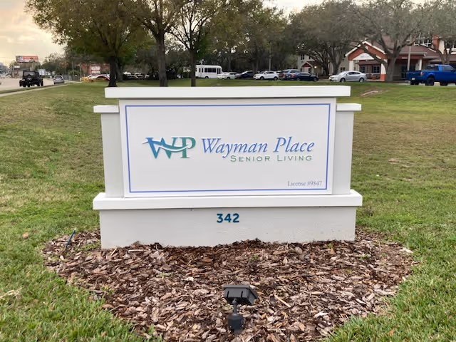 A freestanding lawn sign reading 'Wayman Place Senior Living' with the number 342 in front of grass, trees, and a building.