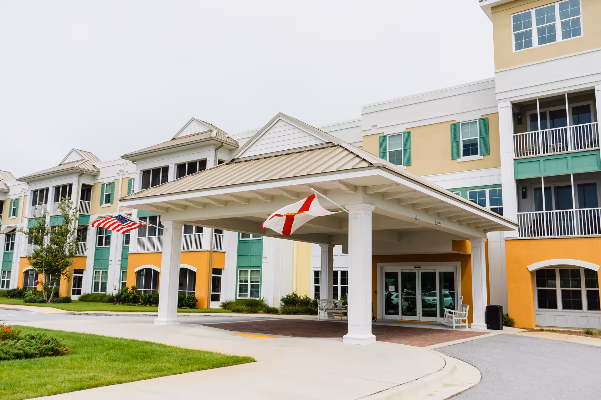 Covered entrance canopy and drop-off area of a colorful multi-story senior living building with American and state flags.