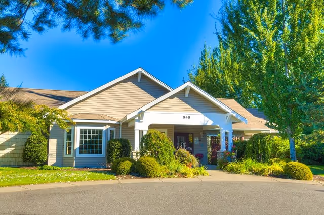 Exterior view of a single-story building with beige siding and a gabled roof, surrounded by well-maintained landscaping including bushes and trees under a clear blue sky.