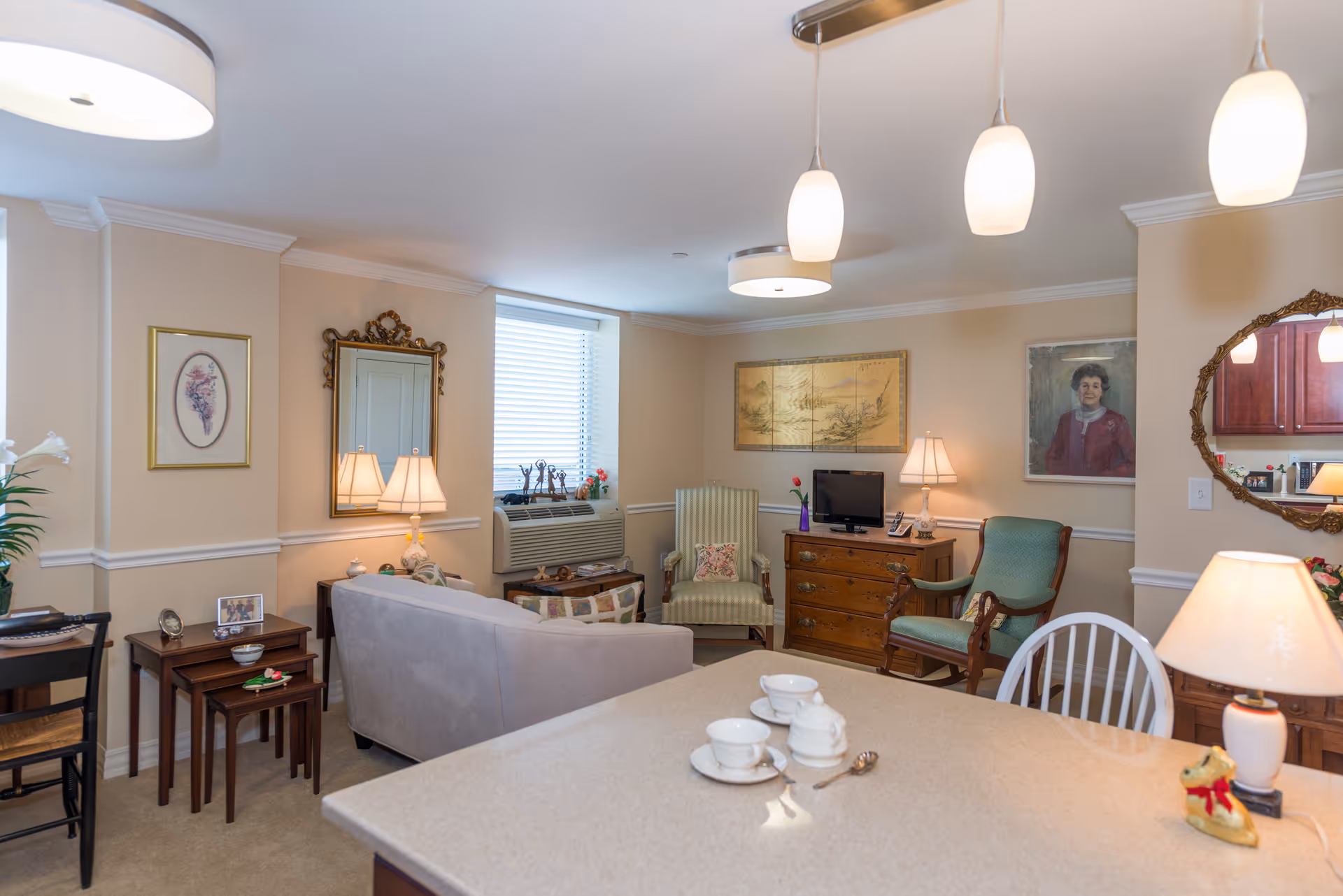 A cozy living room area in a senior living facility with beige walls and carpet. The room features a gray sofa, two armchairs, a wooden dresser with a small TV, and various lamps providing warm lighting. There are framed pictures and a decorative mirror on the walls, a window with blinds, and a countertop in the foreground with white teacups and a teapot.