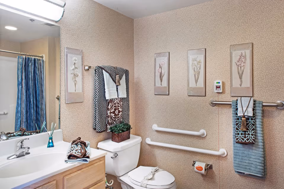 A bathroom with a white sink and wooden cabinet, a toilet with a decorative plant on top, and a shower with a blue curtain. The walls have a beige patterned wallpaper and are decorated with four framed floral prints. There are multiple towels hanging on towel racks, and two white grab bars are installed on the wall near the toilet. A call button is mounted on the wall above the grab bars.