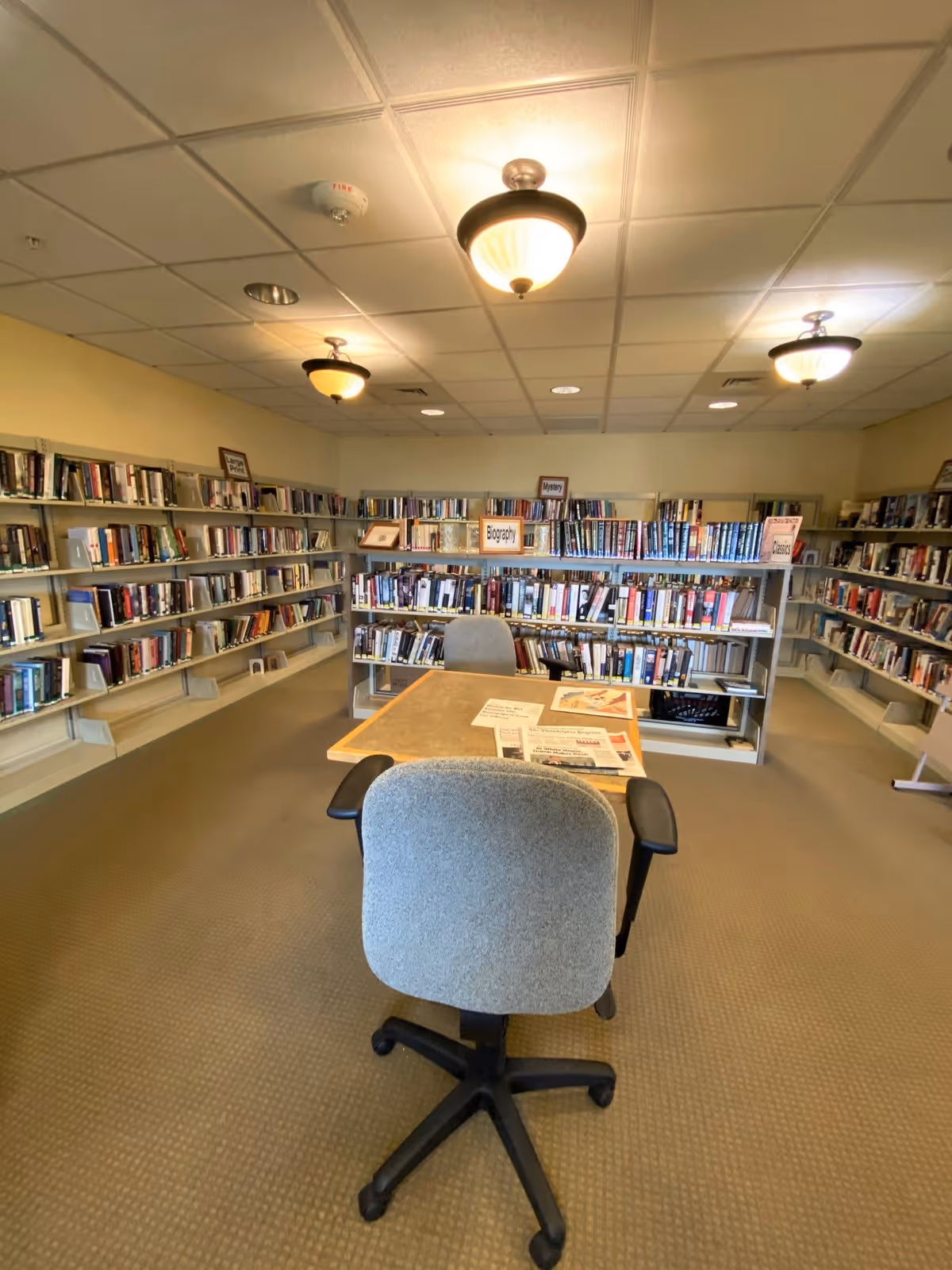 Interior view of a library room with bookshelves lining the walls and a central bookshelf labeled with categories such as Biography, Mystery, and Classics. There is a table with two office chairs, one facing the camera and the other on the opposite side of the table. Papers and newspapers are spread out on the table. The ceiling has recessed lighting and three hanging light fixtures.