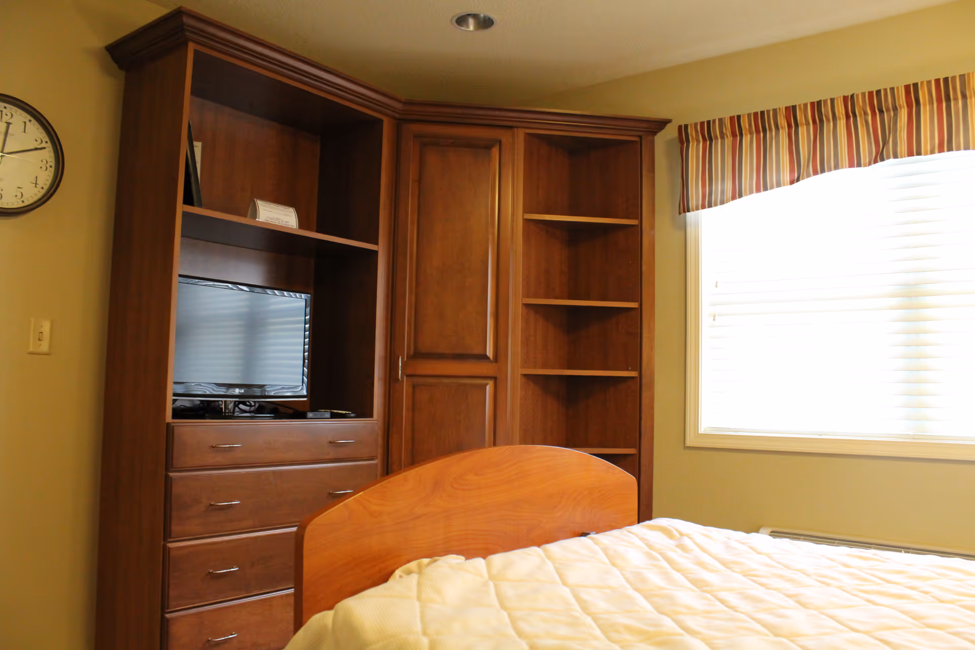 Bedroom with a wooden built-in shelving and dresser holding a small television, a bed in the foreground and a window with blinds.