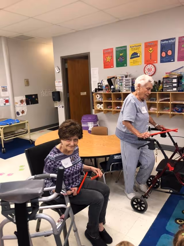 Two elderly women in a room with tables and shelves. One woman is sitting in a chair holding red exercise sticks, and the other woman is standing with a walker, also holding red exercise sticks. The room has colorful posters on the wall and various supplies on shelves.