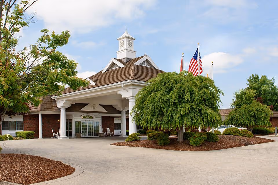 Front entrance of a senior living facility with a covered porte-cochere, manicured trees, and American flags.
