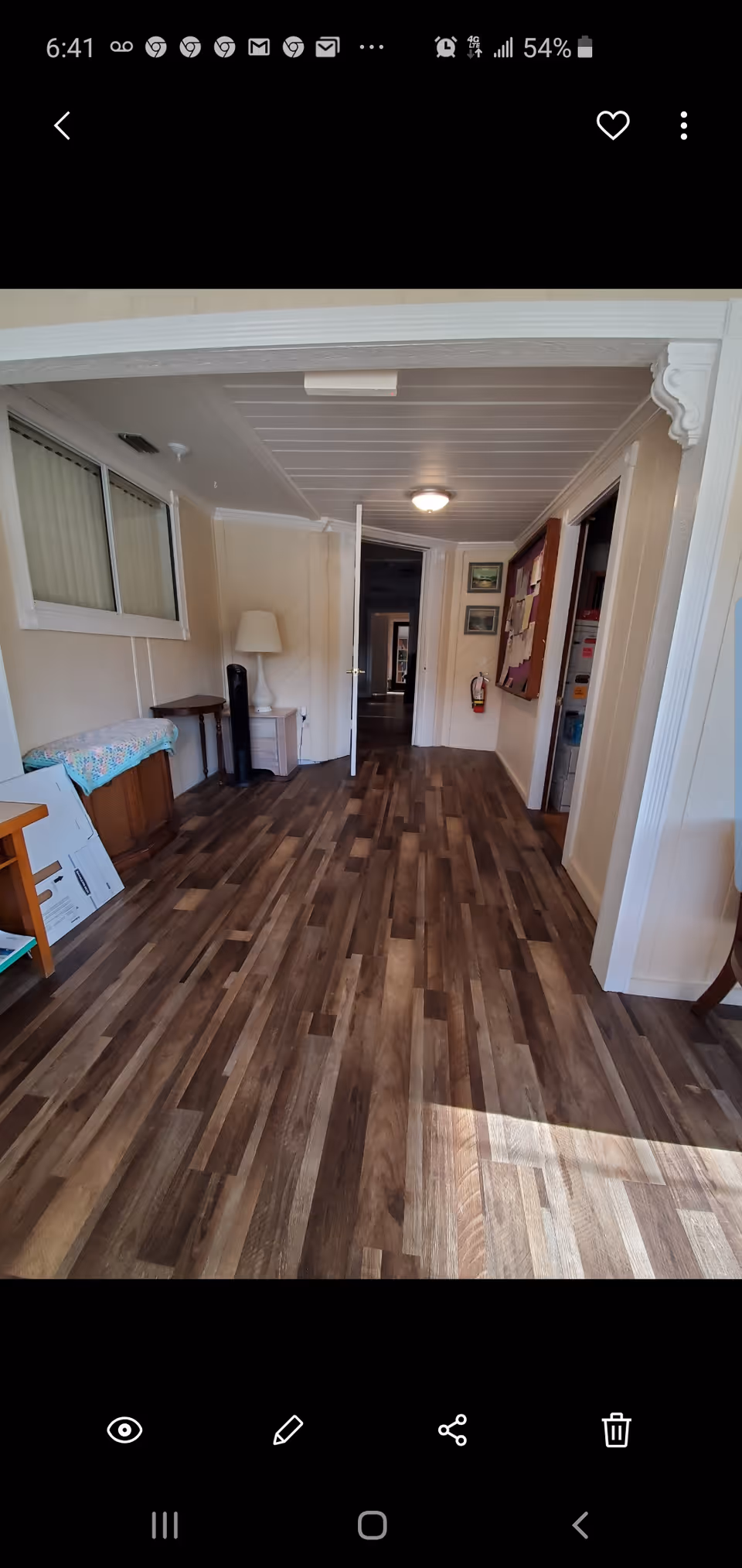 Interior hallway with wood-patterned flooring, beige walls, and white ceiling. There is a white door at the end of the hallway, a bulletin board on the right wall, a fire extinguisher mounted on the wall, and a lamp on a small table to the left. Some furniture and items are placed along the left side of the hallway.