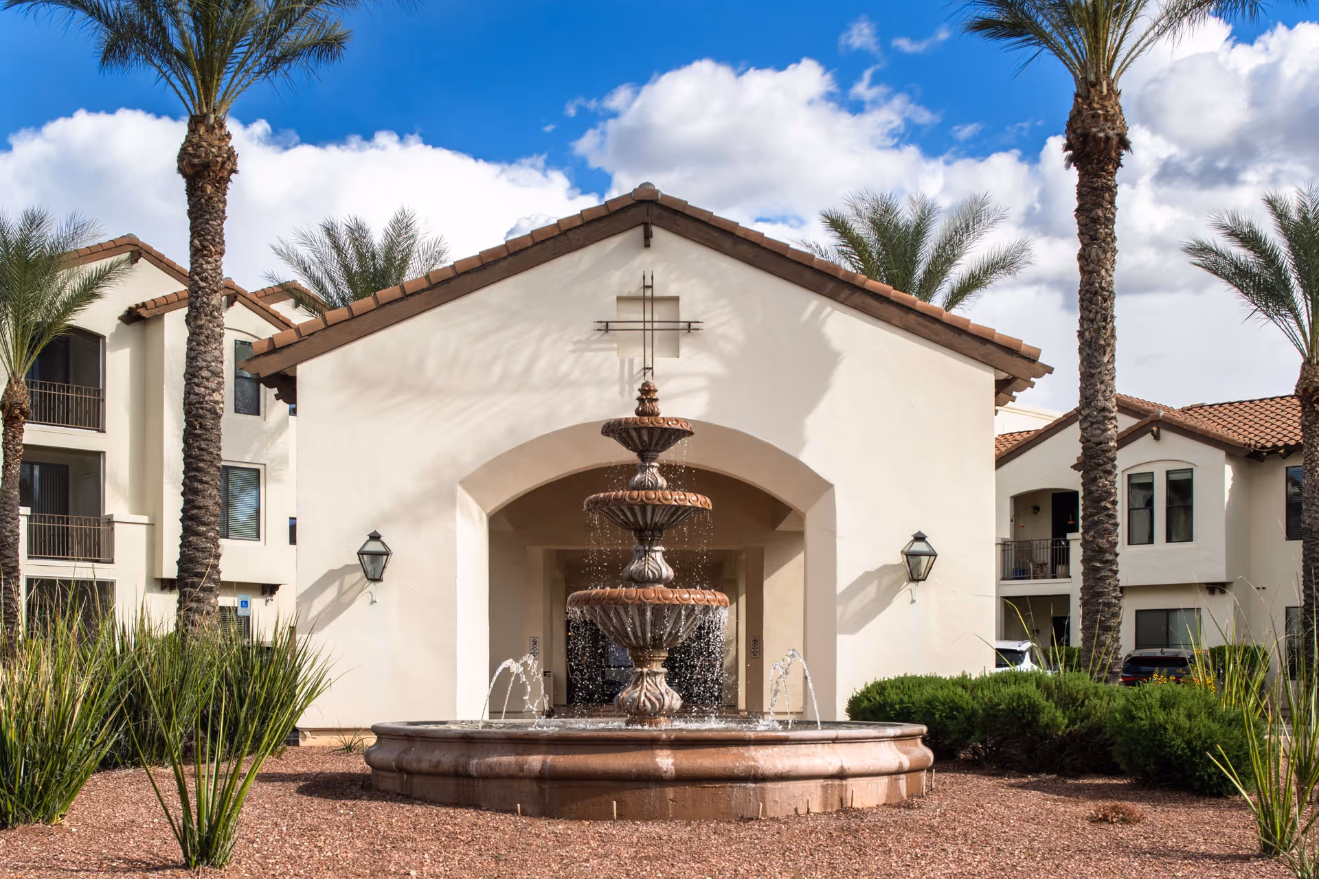 Exterior view of a senior living facility with a three-tiered water fountain in the foreground, surrounded by desert landscaping with palm trees and shrubs. The building has a stucco finish with a tiled roof and balconies.