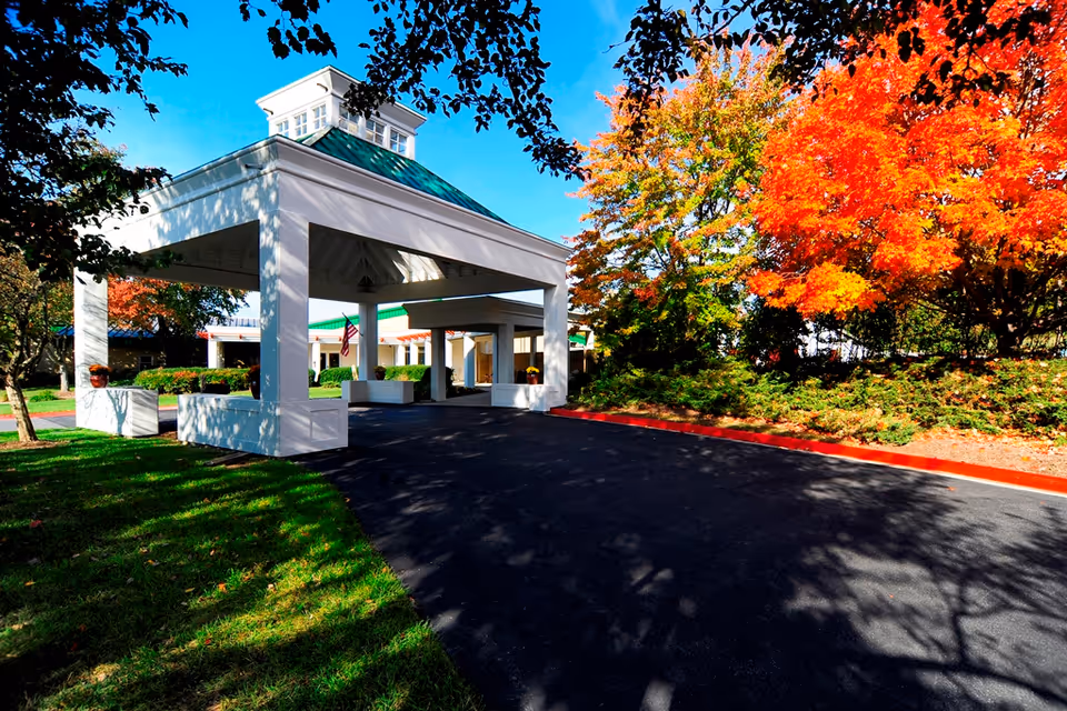 Entrance area of Regency Park Senior Living Community featuring a white covered driveway structure with a green roof, surrounded by green grass and trees with vibrant autumn foliage under a clear blue sky.