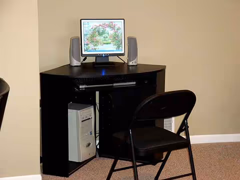 A small computer desk with monitor, speakers, CPU, and a folding chair in a beige room.
