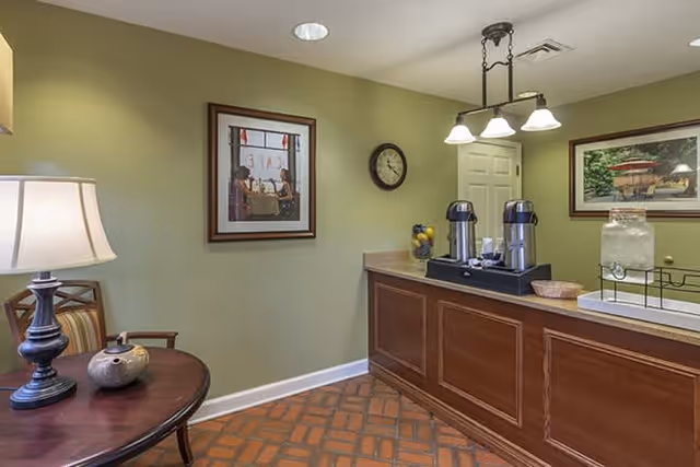 Interior view of a room with a wooden counter holding two coffee dispensers and a glass water dispenser. There is a round table with a lamp and decorative items on the left side. The walls are painted light green and decorated with two framed pictures and a wall clock. The floor has a patterned tile design.