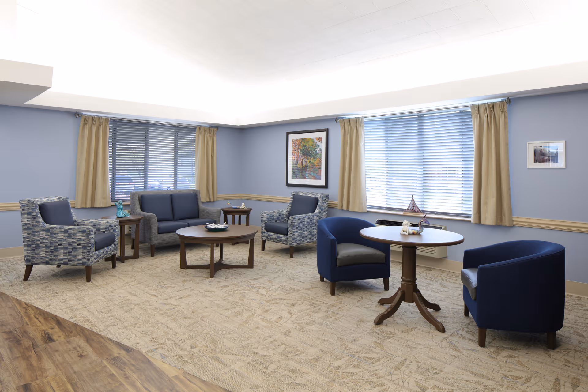 A bright and spacious living room with light blue walls and beige carpet. The room features two large windows with beige curtains and blinds. There are multiple seating options including a blue loveseat, two patterned armchairs, and two solid blue armchairs around a round wooden table. Additional small wooden tables hold decorative items. The room is decorated with framed artwork on the walls.
