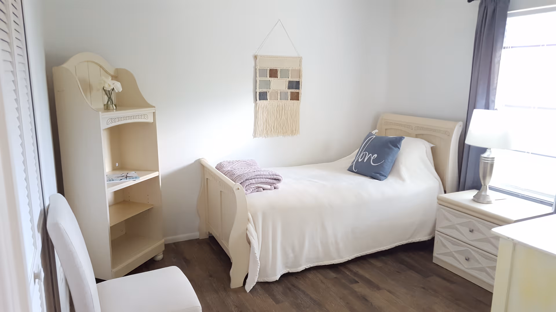A bright, cozy bedroom with a single bed covered in a white blanket and a decorative pillow with the word 'love'. There is a small nightstand with a lamp next to the bed, a white chair, and a small shelving unit with a flower vase on top. A woven wall hanging decorates the white wall above the bed, and a window with gray curtains lets in natural light.