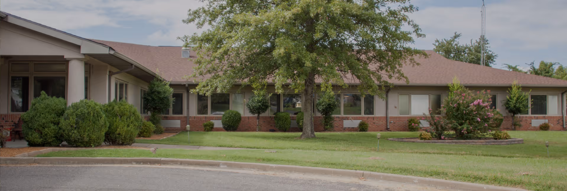 Single-story brick and siding building with a brown roof, surrounded by green grass, bushes, and trees under a partly cloudy sky.