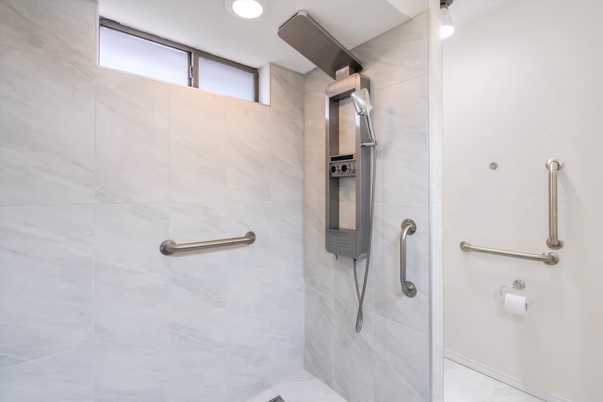 Accessible tiled shower with overhead and handheld shower fixtures, stainless grab bars, and a wall-mounted toilet paper holder.