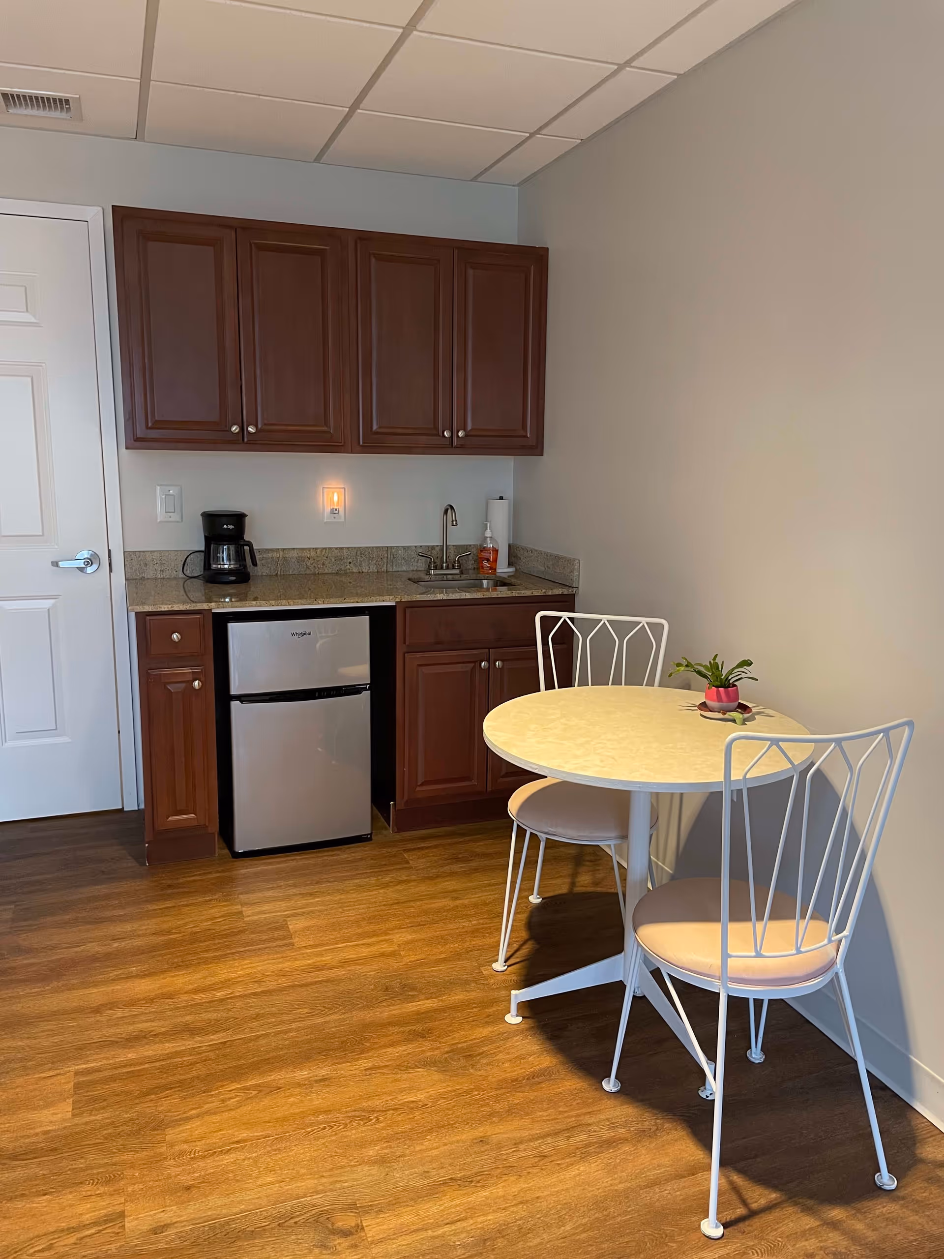 Small kitchenette area with wooden cabinets, a mini refrigerator, a coffee maker, a sink, and a round table with two white metal chairs with light-colored cushions. The floor is wooden, and the walls are light-colored.