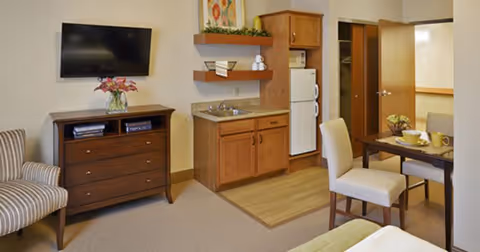 Interior view of a senior living facility room featuring a small kitchenette with wooden cabinets, a refrigerator, and a sink. There is a dining table with two chairs set with dishes and cups. A flat-screen TV is mounted on the wall above a wooden dresser, and a striped armchair is positioned nearby. The room has a neutral color palette with beige walls and carpeted flooring.