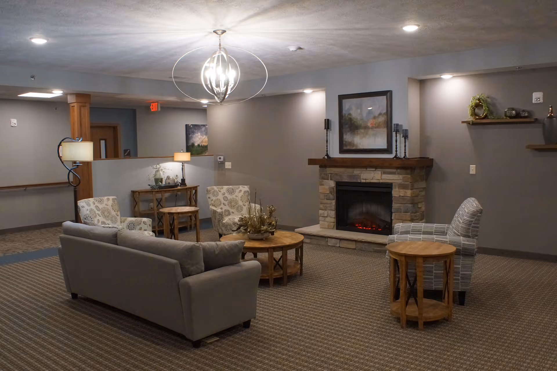 A cozy common area with a gray sofa, two patterned armchairs, and a striped armchair arranged around a round wooden coffee table and a smaller side table. The room features a stone fireplace with a painting above it, wall shelves with decorative items, and soft lighting from ceiling fixtures and lamps. The carpet is patterned, and the walls are painted in neutral tones.