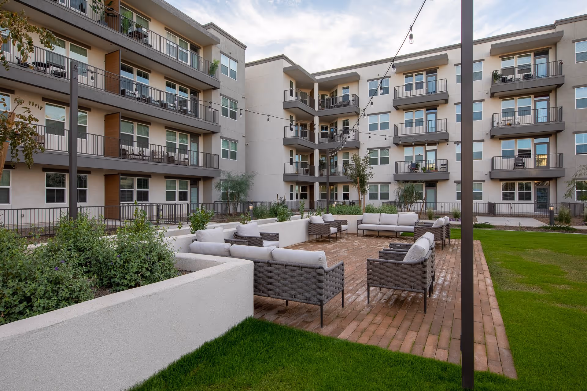 Outdoor courtyard area of a multi-story residential building with balconies. The courtyard features a seating area with cushioned chairs and sofas arranged on a brick patio, surrounded by green grass and plants. String lights are hung above the seating area.