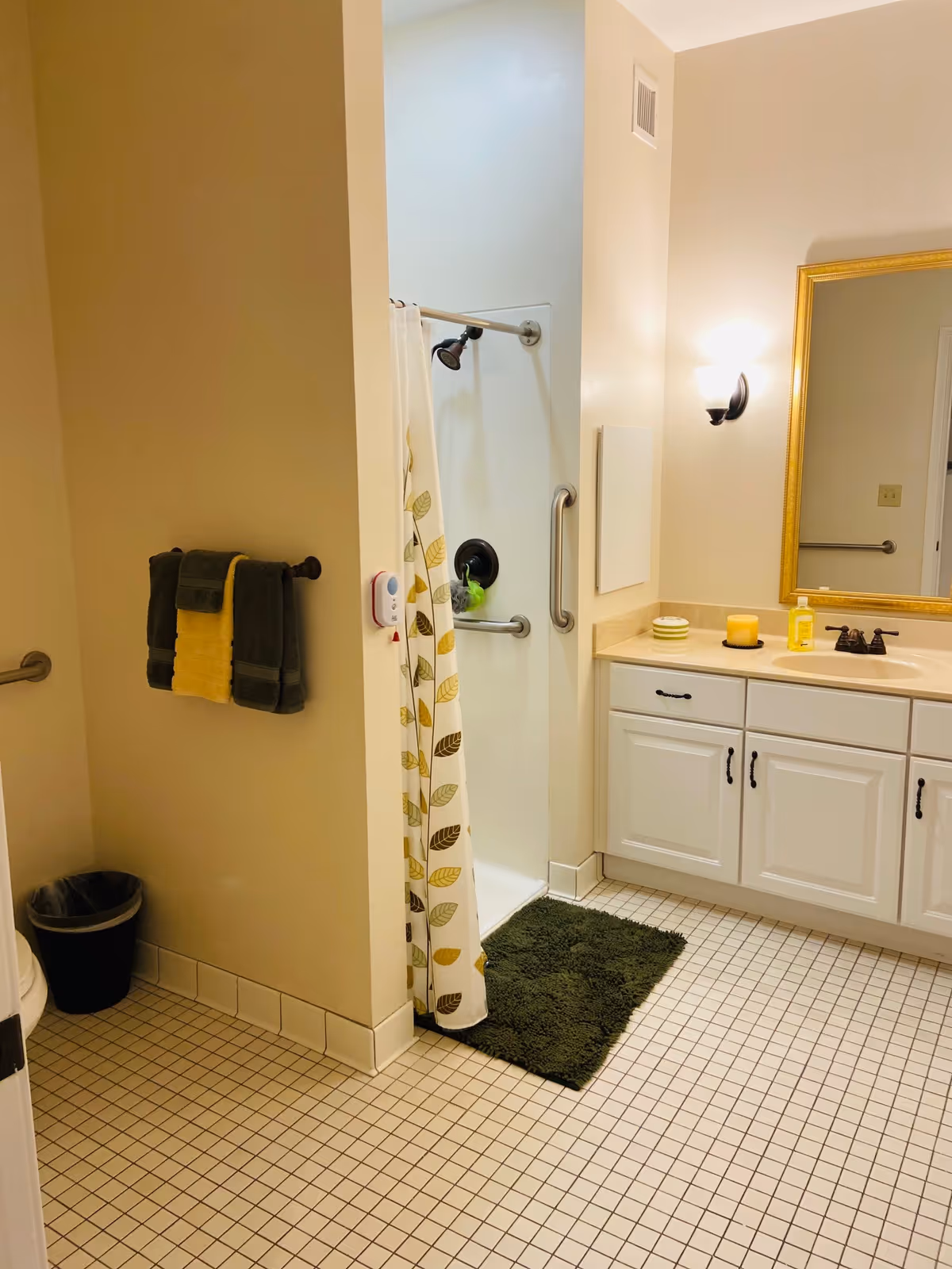 A clean bathroom with a tiled floor, a walk-in shower with a curtain featuring a leaf pattern, a green bath mat, a white vanity with a sink, a large gold-framed mirror, and a wall-mounted light fixture. Towels are hanging on a rack next to the shower, and a small trash can is visible near the toilet area.