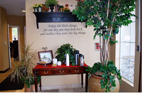 Interior corner of a senior living facility with a wooden table holding coffee supplies, cups, and a framed certificate. Above the table is a black wall shelf with decorative items and a wall decal that reads, 'Enjoy the little things, for one day you may look back and realize they were the big things.' There are several potted plants around the table, including a large leafy plant in a large pot to the right near a window.