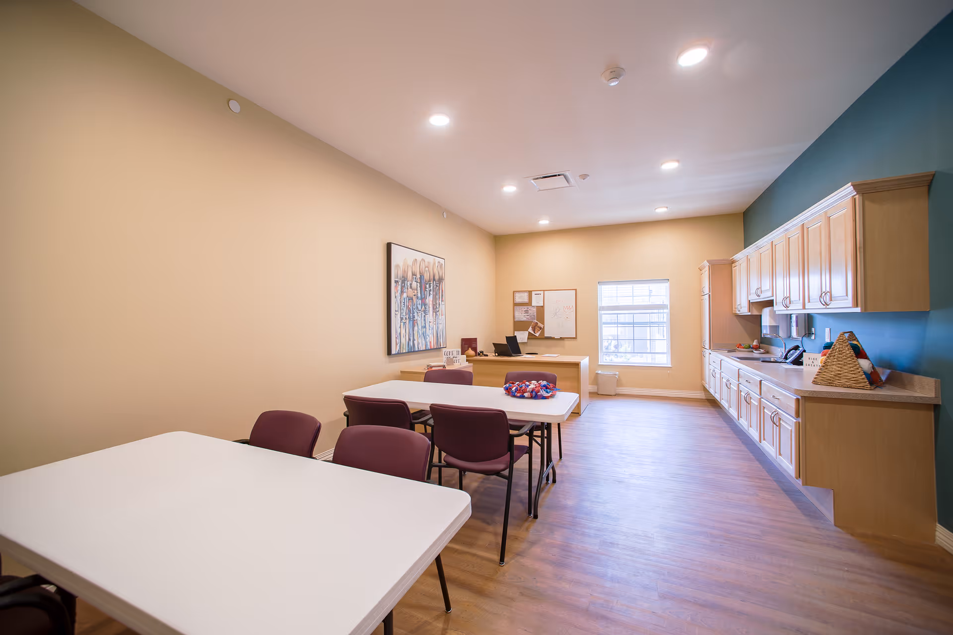 A bright room with beige walls and wood flooring featuring two white tables with purple chairs around them. On the right side, there is a long countertop with wooden cabinets above and below. At the far end of the room, there is a desk with a computer and office supplies, a window letting in natural light, and a bulletin board on the wall.