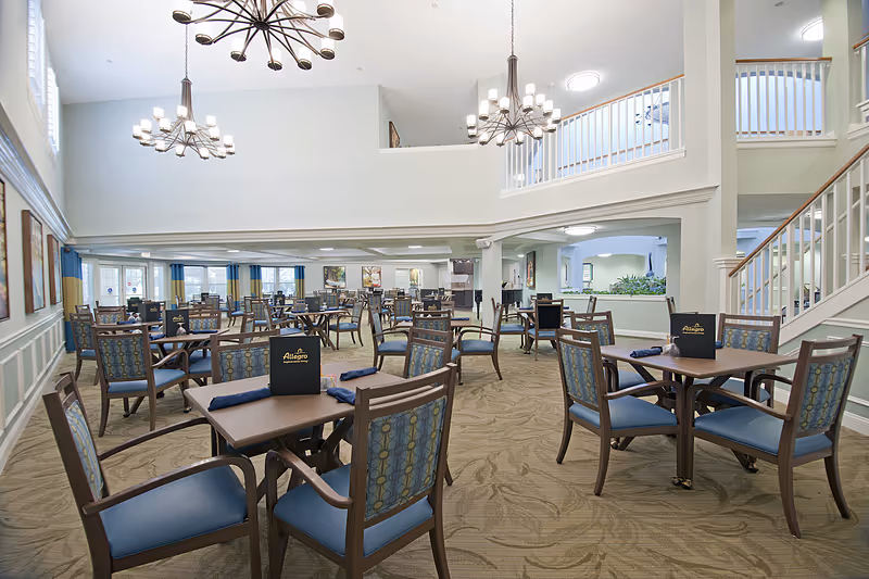 Spacious dining room in Allegro Senior Living – Fleming Island, FL with multiple wooden tables and blue cushioned chairs arranged neatly. The room features high ceilings with modern chandeliers, large windows allowing natural light, and a staircase leading to an upper level with white railings.