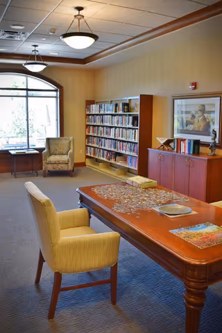 A cozy reading room with a large wooden table in the center covered with a partially completed jigsaw puzzle. Two upholstered chairs are positioned around the table. In the background, there is a bookshelf filled with books, a wooden cabinet with decorative items and framed artwork above it, and a large window letting in natural light.