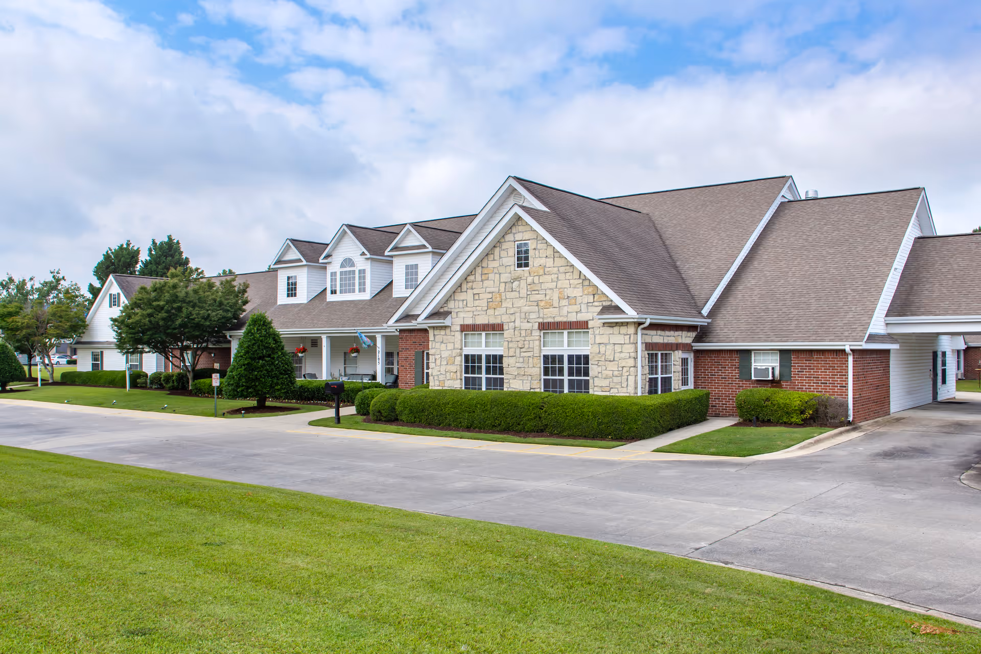 Exterior view of a single-story senior living facility building with a combination of stone, brick, and white siding. The building has multiple gabled roofs and several windows. There is a well-maintained lawn, trimmed bushes, and a paved driveway in front of the building under a partly cloudy sky.