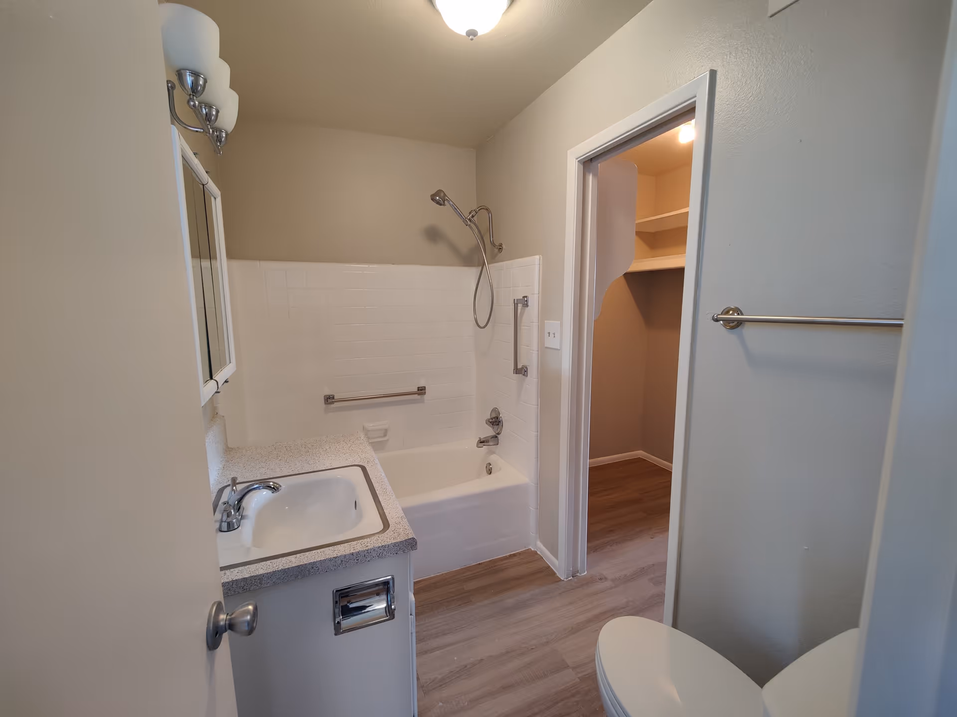 Interior view of a bathroom featuring a white bathtub with a showerhead and grab bars, a sink with a countertop and faucet, a mirror with lights above it, a towel rack on the wall, and an open doorway leading to a closet with shelves and wooden flooring.