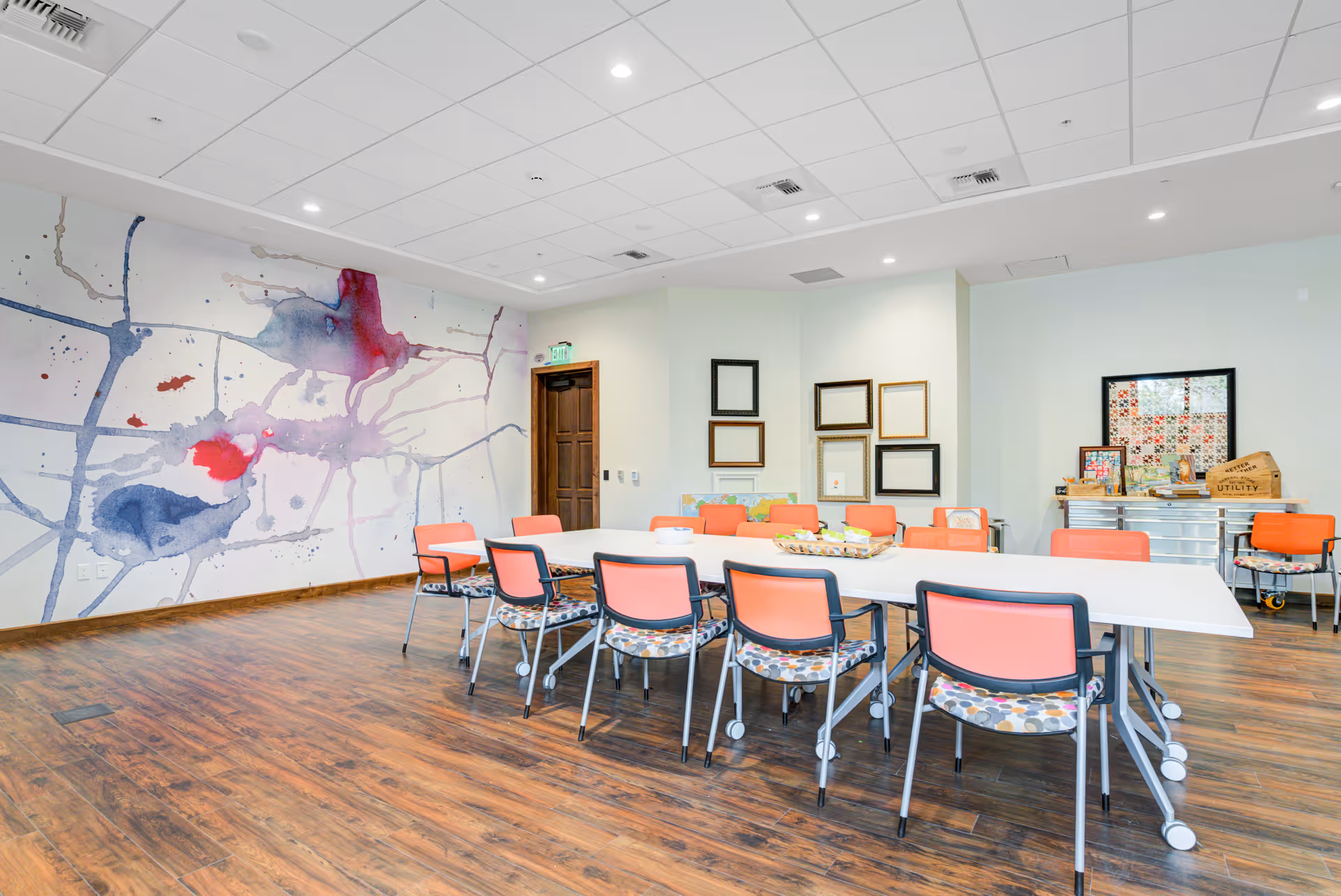 A brightly lit room with a large white table surrounded by orange and patterned chairs on wheels. The room has wooden flooring and a white ceiling with recessed lighting. One wall features an abstract watercolor mural with splashes of blue, purple, and red. Another wall has several empty picture frames arranged in a grid, and a cabinet with decorative items and a framed quilt is visible.