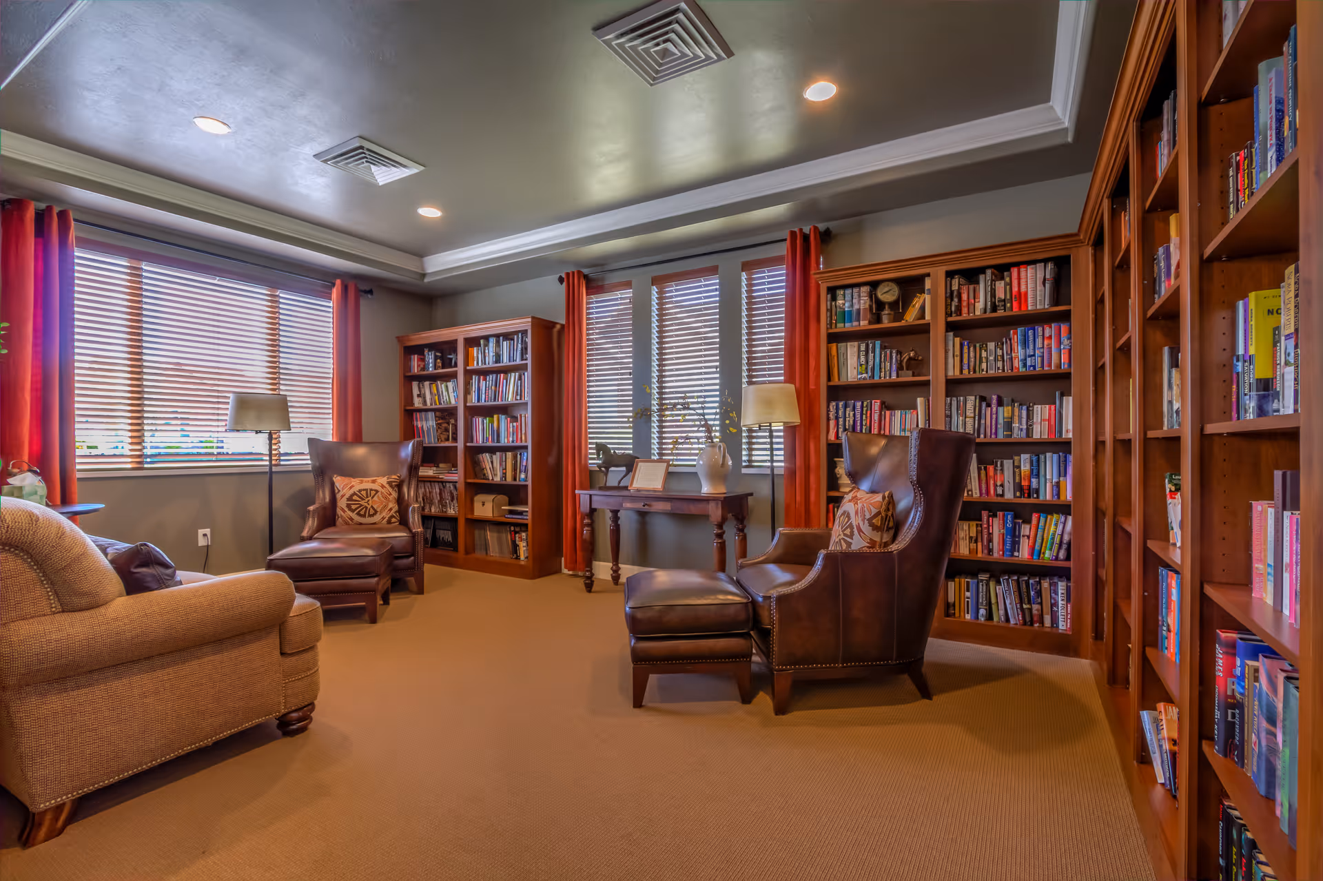 A cozy library room with large wooden bookshelves filled with books, two leather armchairs with matching ottomans, a beige sofa, a wooden table with a lamp and decorative items, and windows with wooden blinds and red curtains.