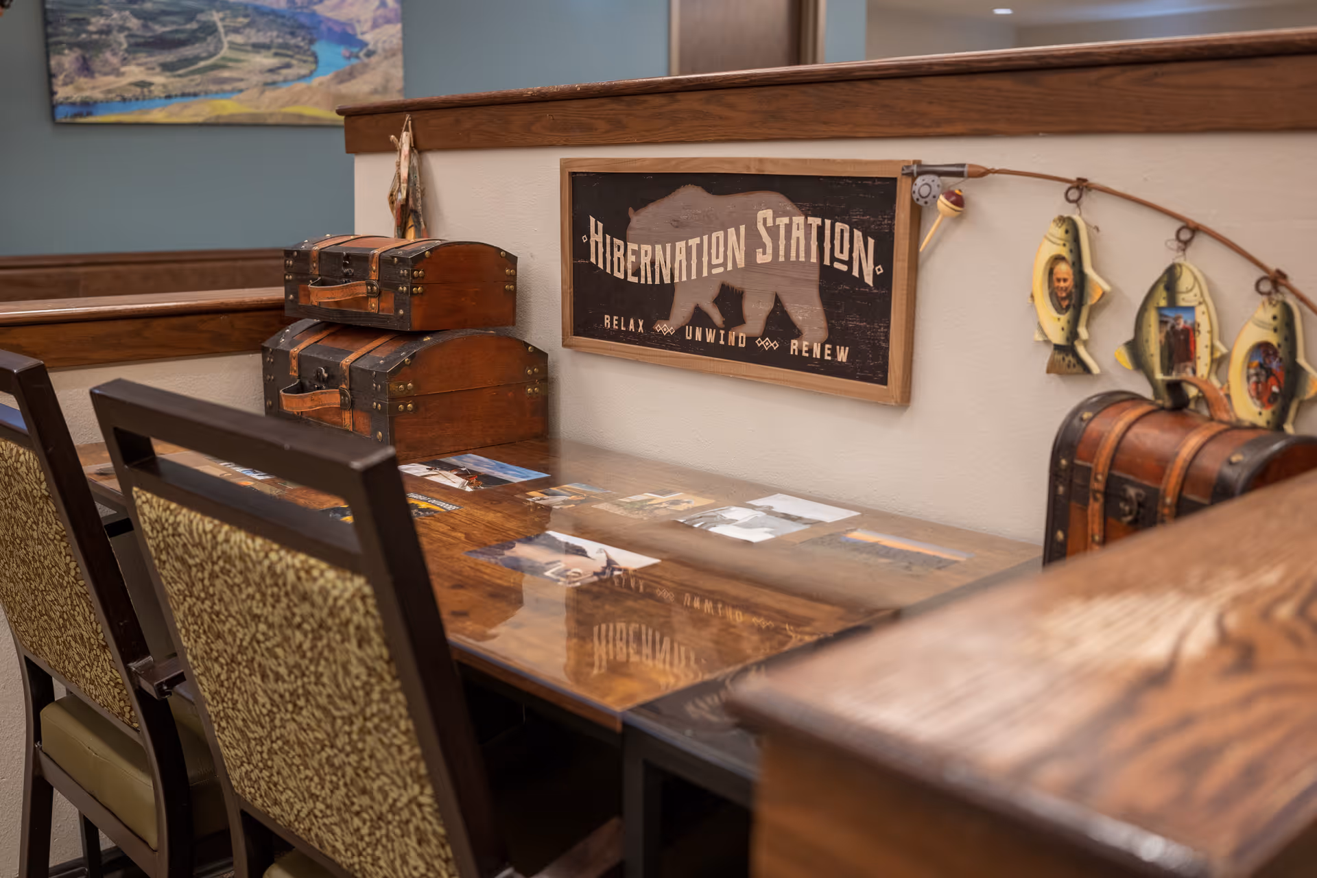 A cozy interior corner with a wooden table and two patterned chairs. On the table are two stacked wooden chests and several photographs under a glass surface. Above the table, a wall sign reads 'Hibernation Station - Relax Unwind Renew' with a bear silhouette. To the right, a fishing rod is mounted on the wall with three fish-shaped photo frames hanging from it. A landscape painting is partially visible on the blue wall in the background.