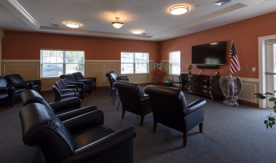 A common area in an assisted living facility with multiple black leather armchairs arranged in rows facing a wall-mounted flat screen TV. The room has three windows with blinds, a popcorn machine in the corner, an American flag, and a decorative eagle statue near the TV stand. The walls are painted a warm reddish-brown color with beige wainscoting, and the ceiling has several round light fixtures.
