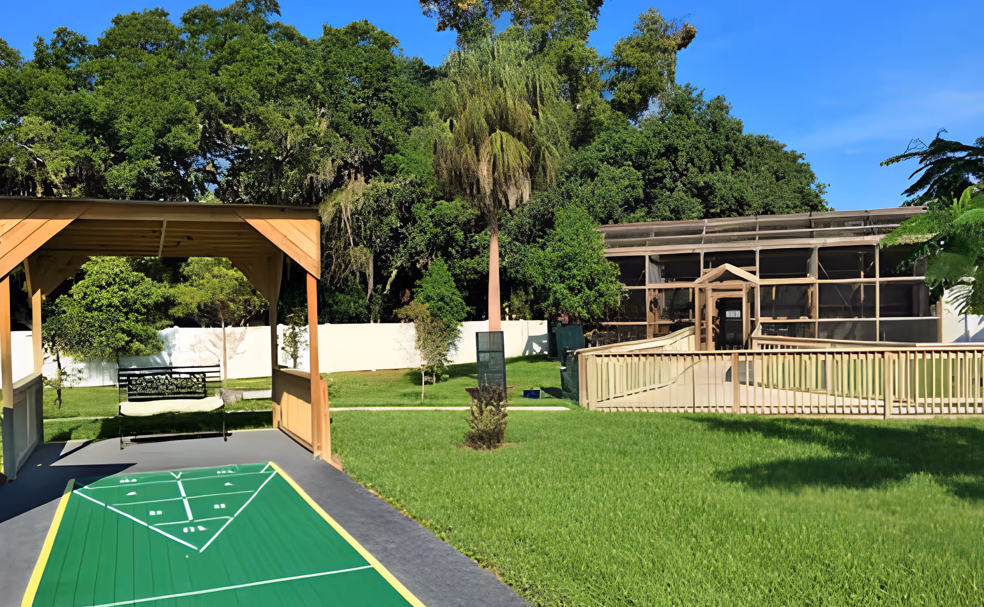 Outdoor courtyard featuring a covered shuffleboard area, a bench swing, grassy lawn, and a screened enclosure building with trees in the background.