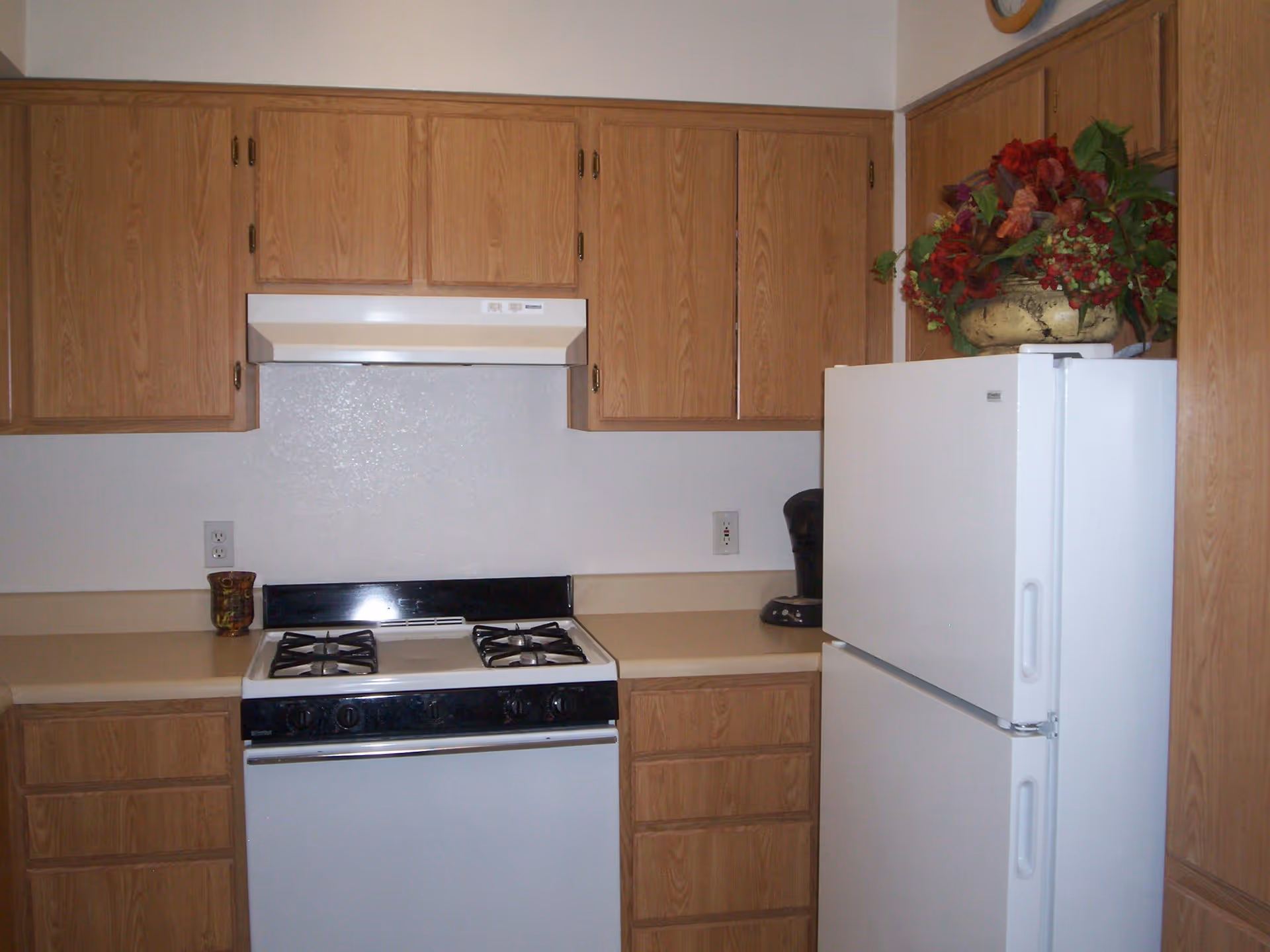 A kitchen with wooden cabinets, a white gas stove with four burners, a white range hood, a white refrigerator, a coffee maker on the countertop, and a decorative flower arrangement on top of the refrigerator.