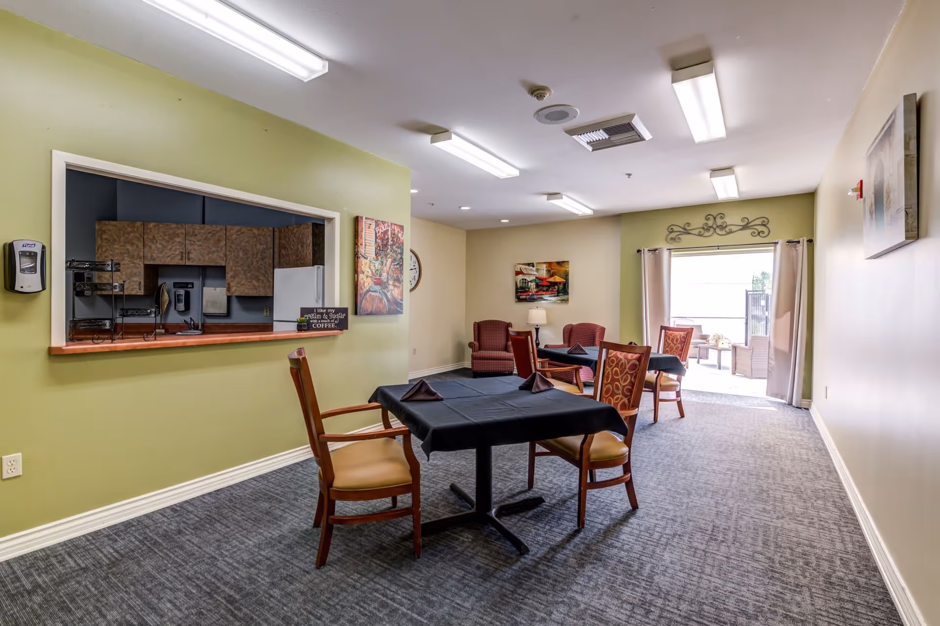 Communal dining and lounge area with tables and chairs, a kitchen pass-through counter, and a doorway to an outdoor patio.