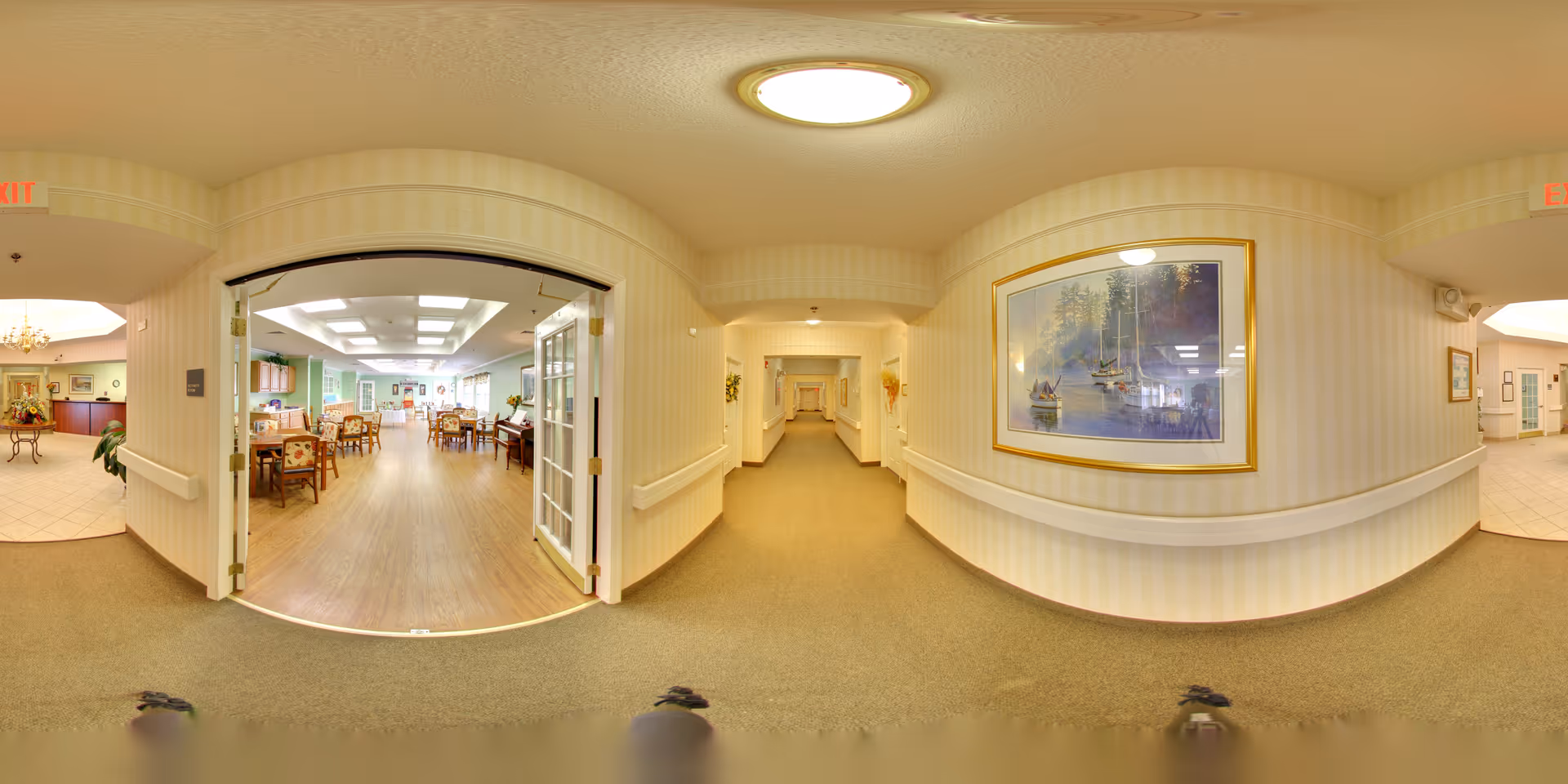 A wide-angle view of a hallway in a senior living facility with beige walls and carpet. On the left, double glass doors open into a dining area with wooden tables and chairs, and a piano. The hallway extends straight ahead with framed artwork on the walls. Ceiling lights illuminate the space.