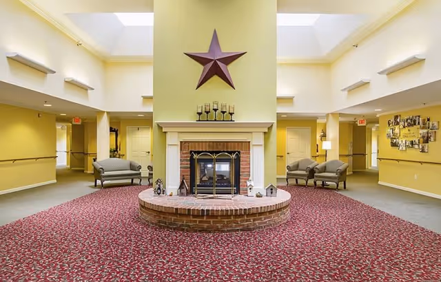 A spacious common area with a central brick fireplace topped by a large decorative star. The room has yellow walls, a patterned red carpet, and several seating areas with armchairs. There are skylights in the ceiling allowing natural light to brighten the space, and hallways leading off the room.