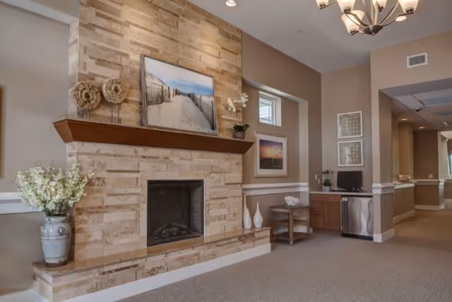 A cozy interior common area with a stone fireplace, a wooden mantel displaying decorative items and a framed beach photograph. To the right, there is a small desk with a computer and a mini refrigerator underneath. The walls are painted in neutral tones with framed artwork and a vase with flowers on a small table. A chandelier hangs from the ceiling.